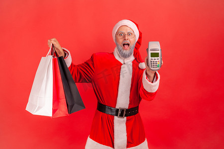 圣诞老人白胡子摄影照片_兴奋的白胡子老人穿着圣诞老人服装，手里拿着购物袋，向镜头展示终端，进行非接触式支付。