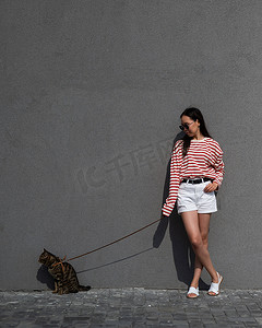 一位年轻女子牵着一只灰色虎斑猫，拴在灰色的墙上。