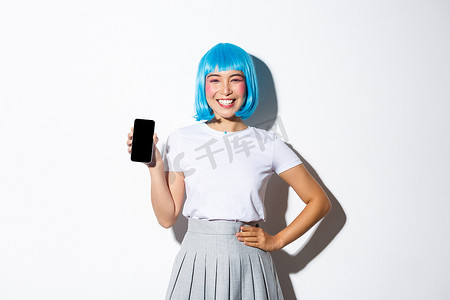 可爱的亚洲女孩的肖像，穿着蓝色假发的动漫人物，微笑着，展示智能手机屏幕，站在白色背景上