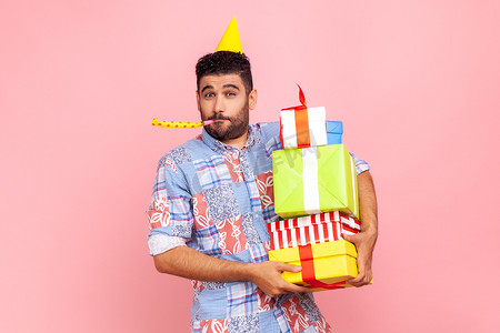 快乐有趣的男人的肖像，留着胡子，穿着休闲风格的衬衫和锥体，拿着一堆礼物，吹着派对号角，庆祝生日。