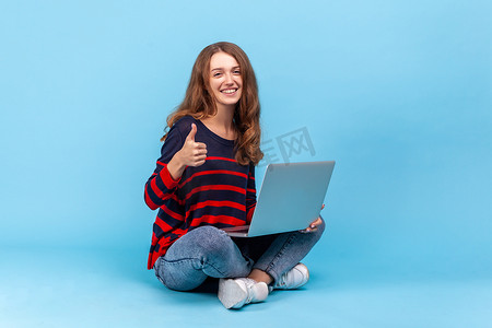 女人坐在地板上，笔记本电脑放在地上，在线工作，竖起大拇指，对远程办公感到满意。