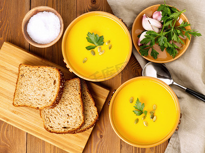 木质盘子和勺子摄影照片_两碗棕色南瓜汤，配欧芹和南瓜籽，棕色深色木质背景，顶视图