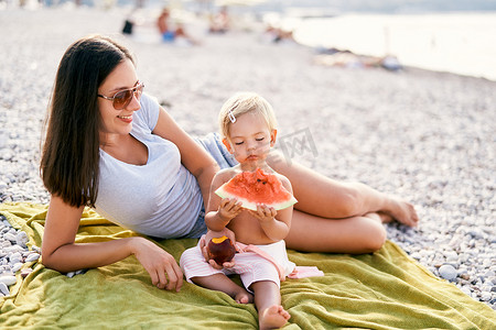 微笑的妈妈向在海滩上吃西瓜的小女孩递桃子