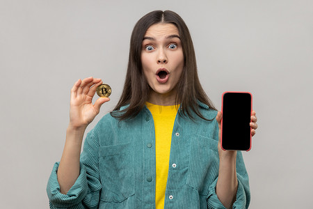 惊讶的女人拿着黄金比特币和智能手机，屏幕空空，用于广告或促销。
