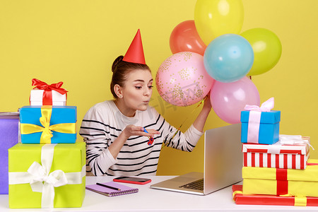 坐在工作场所的女性被礼品盒和气球包围，向笔记本电脑屏幕发送飞吻。
