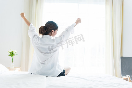 女人早上在靠近窗户的床上醒来后伸展身体的后视图。
