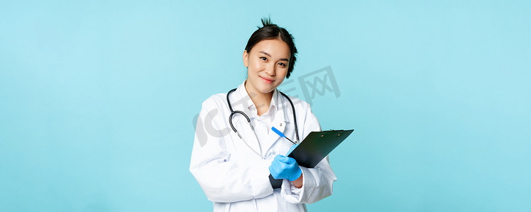 女医生、身穿医疗制服的亚洲医生的肖像，写下患者信息，站在蓝色背景下