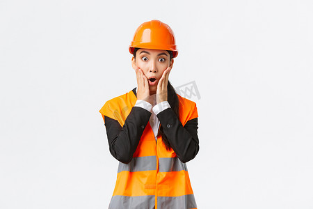 女脸手摄影照片_震惊而担忧的亚洲女工程师在建筑区域遇到问题，盯着项目，惊慌失措的脸，手握在嘴边，担心地喘着气，站在白色背景上