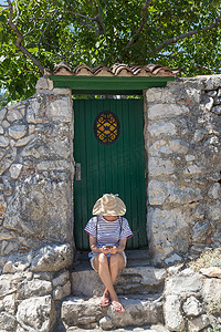 美丽的年轻女游客戴着大草帽，使用莫伊莱电话，坐在地中海老城的白色木门和纹理石墙前