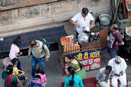 空中无人机拍摄的路边茶贩穿着白衬衫库尔塔，小摊位在炉子和旧器皿上准备茶，在繁忙的城市里，人群在他周围走动，放入牛奶、树叶、糖等