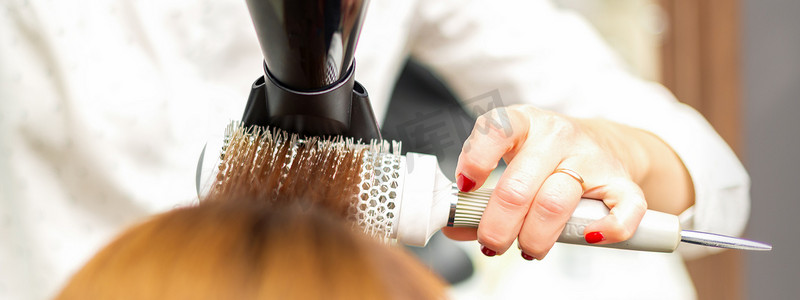 一位专业美发师正在用吹风机和圆刷烘干长红头发，特写。