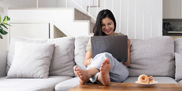 亚洲年轻女性看着笔记本电脑，笑着说好消息或在家网上购物的折扣券。看着笔记本电脑的快乐而开朗的女性读到消息感到兴奋
