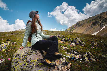 坐在石头摄影照片_一位背着背包的年轻女子坐在石头上，看着天空中的云彩和山脉，远足度假