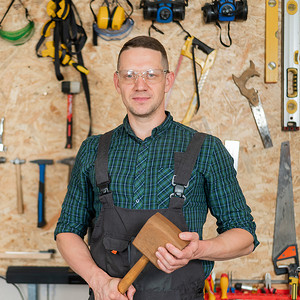 一位戴着护目镜和工作服的木匠在车间里拿着一把木锤的肖像，背景是墙上有工具。