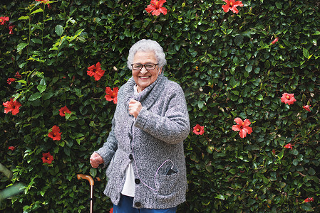 有趣的老妇人戴着耳机在智能手机上跳舞听音乐，微笑着在带花墙的花园里享受庆祝退休的乐趣