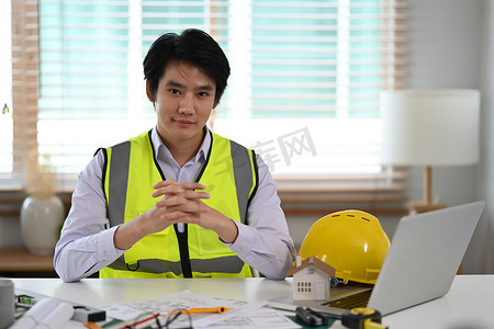 身穿白衬衫和黄色背心的英俊男性建筑工程师坐在工作场所，对着镜头微笑。