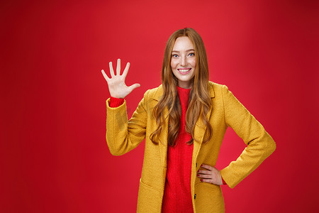给星摄影照片_女孩给很棒的网上商店打了五颗星，她买了一套适合秋季天气的衣服，举起的手掌微笑着，站在红色背景的时尚温暖的黄色外套上
