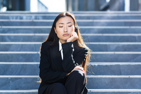 怀孕悲伤摄影照片_年轻美丽的亚洲女性坐在办公中心的楼梯上，心烦意乱、悲伤