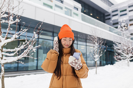 走路听音乐摄影照片_年轻美丽的亚洲女性在冬雪天走路时用耳机听音乐，使用手机在线应用程序