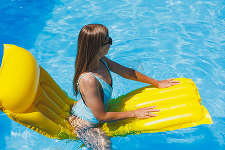 身穿泳衣、戴着太阳镜的漂亮年轻女子躺在泳池里的黄色充气床垫上。