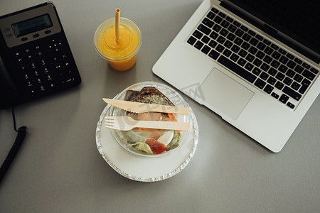 办公室工作场所的平躺、盒中午餐和桌上的橙汁饮料，配有笔记本电脑和电话