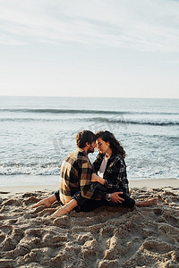 海边女子摄影照片_日出时，年轻女子和男子坐在海边拥抱，背景是海浪