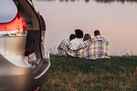 湖边摄影照片_日落时分，四口之家坐在湖边的草地上，父母带着两个孩子享受周末公路旅行