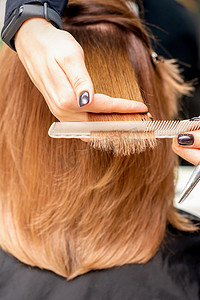 美发师的手将发束夹在手指之间，在美发沙龙里用梳子和剪刀为年轻女子的长发进行理发，特写。