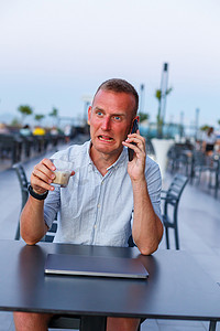 商人在酒店的露台上使用笔记本电脑并通过电话交谈，这是在度假村度假期间管理员工的时间。