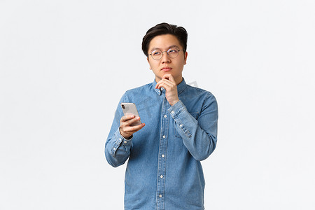 戴着眼镜、有思想、有创意的亚洲男人在社交媒体上发帖时思考，把目光移开，思考或做出决定，拿着智能手机，在互联网上选择一些东西