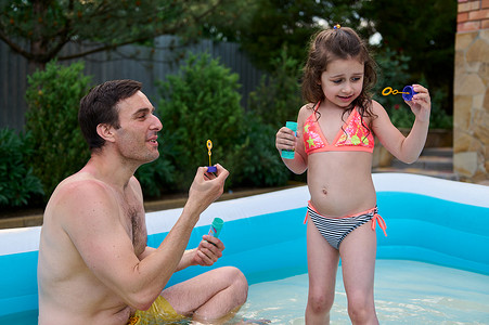 穿着泳衣的可爱小女孩在她慈爱的爸爸旁边的充气池里吹肥皂泡。