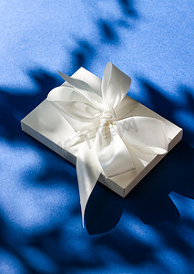 白色蝴蝶结摄影照片_蓝色背景上带丝带和蝴蝶结的豪华假日白色礼盒、豪华婚礼或生日礼物