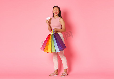 乐观、兴奋、微笑的时尚亚洲女孩的全长肖像，展示她的信用卡，看上去很高兴，拎着购物袋，买了度假新衣服，粉红色背景