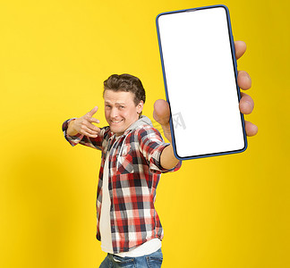 很高兴赢得英俊的男子指着白色空屏幕的智能手机，穿着红色格子衬衫和牛仔裤，手机显示屏在黄色背景上隔离。