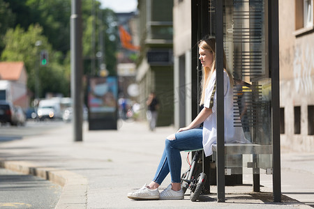 休闲白人青少年通勤者，拥有现代可折叠城市电动滑板车，坐在公交车站长凳上等待地铁城市公交车。