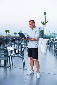 一位成功的商人在酒店露台上使用笔记本电脑工作，并在异国情调的度假胜地度假时抽出时间远程管理员工。