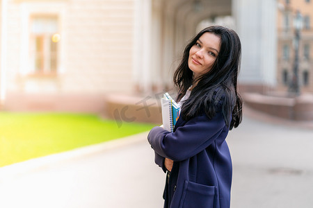 美丽的微笑女孩黑发学生拿着笔记本和课本，站在圣彼得堡街上的大学。