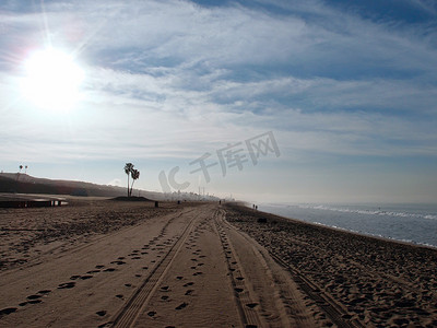 沙子上的轮胎痕迹和脚印，远处有棕榈树和发电厂，太阳升起