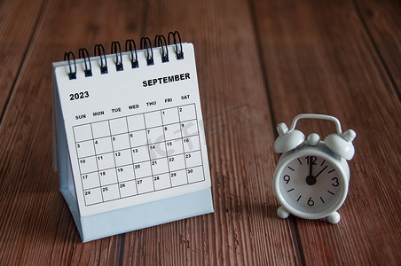 闹钟列表摄影照片_2023 年 9 月白色台历，闹钟指向木桌上 12 点。