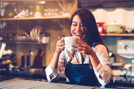 快乐的亚洲女咖啡师拿着一杯咖啡，品尝自己煮的咖啡，有咖啡馆餐厅的背景。