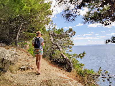 偏远的摄影照片_年轻活跃的女性游客穿着小背包，在松树间的沿海小路上行走，寻找偏远的海湾，在克罗地亚的海边和平地独自游泳。