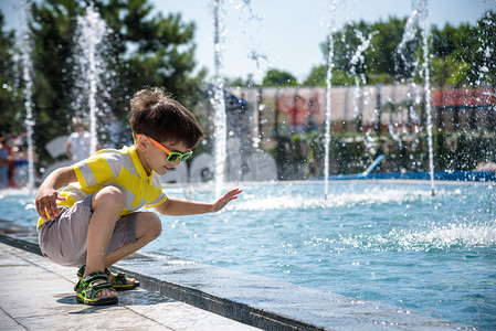 阳光明媚的夏日，小男孩在游泳池附近的广场上玩耍，喷泉里有喷水。