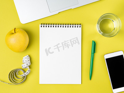 空白笔记本页，用于饮食计划或菜单、卷尺、玻璃水和亮黄色背景的苹果、减肥概念、顶视图。