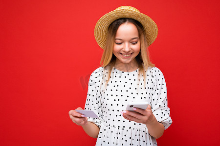 照片中，身穿夏装、开朗漂亮的金发女郎使用手机，手持信用卡在红色背景下在线支付。