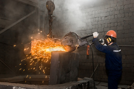 一名戴着防护头盔、呼吸器、工作服的男工人在工厂车间管理带有飞火花的铸铁混凝土管的重型研磨设备