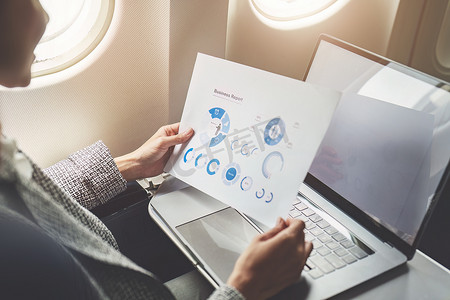 一位成功的亚洲女商人或女性企业家在飞机上穿着正装，坐在商务舱座位上，在飞行过程中使用带有笔记本电脑的文件