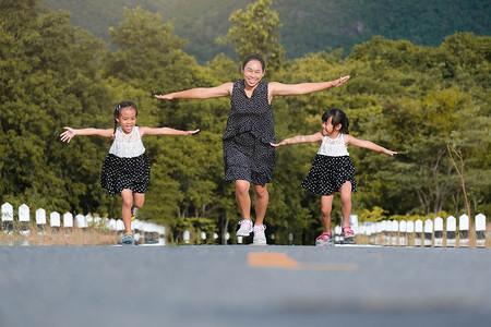 两个可爱的女儿在妈妈张开双臂的陪伴下，在公园的马路上奔跑，仿佛飞翔。