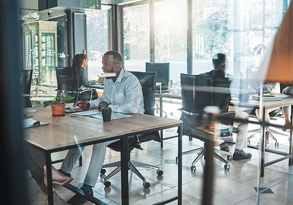 商务、办公室和工作，一个男人坐在玻璃大楼的办公桌前，与后台的同事一起工作。