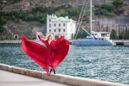 一个穿着红色连衣裙的女人，一个穿着飘逸连衣裙、长着长丝翅膀的时装模特，堤岸上飞扬着织物。