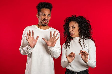 不满摄影照片_年轻的非洲夫妇站在红色工作室背景上表达不满，并对镜头表现出厌恶、厌恶的姿态。